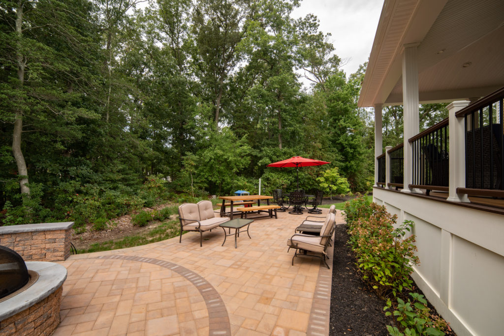 A brick patio with outdoor furniture and firepit features a dynamic design, flanked with lush green trees and landscaping.
