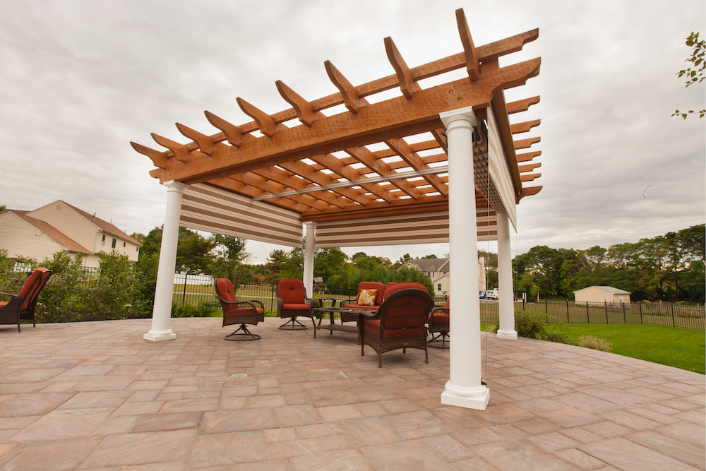 Brown pergola on brick floor in backyard, with 2 red chairs underneath. 
