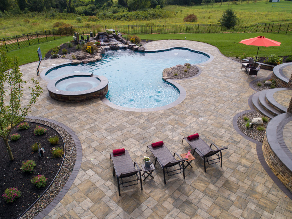 Freeform pool featuring a waterfall and inbuilt spa, with a stone patio and three lounge chairs.