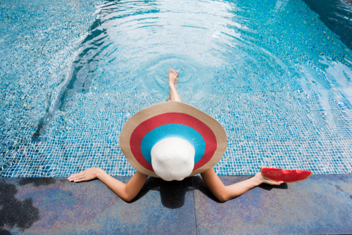 Woman with a white, blue and red sombrero relaxes as she puts her legs into a pool with a shimmering tile interior. 
