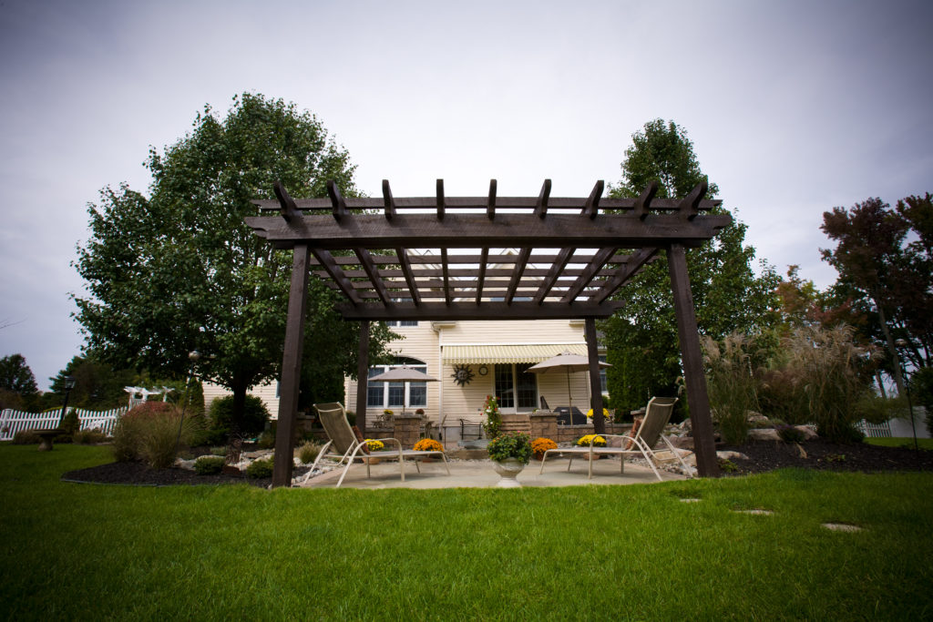 Dark brown pergola with two lounge chairs underneath it in the backyard. 