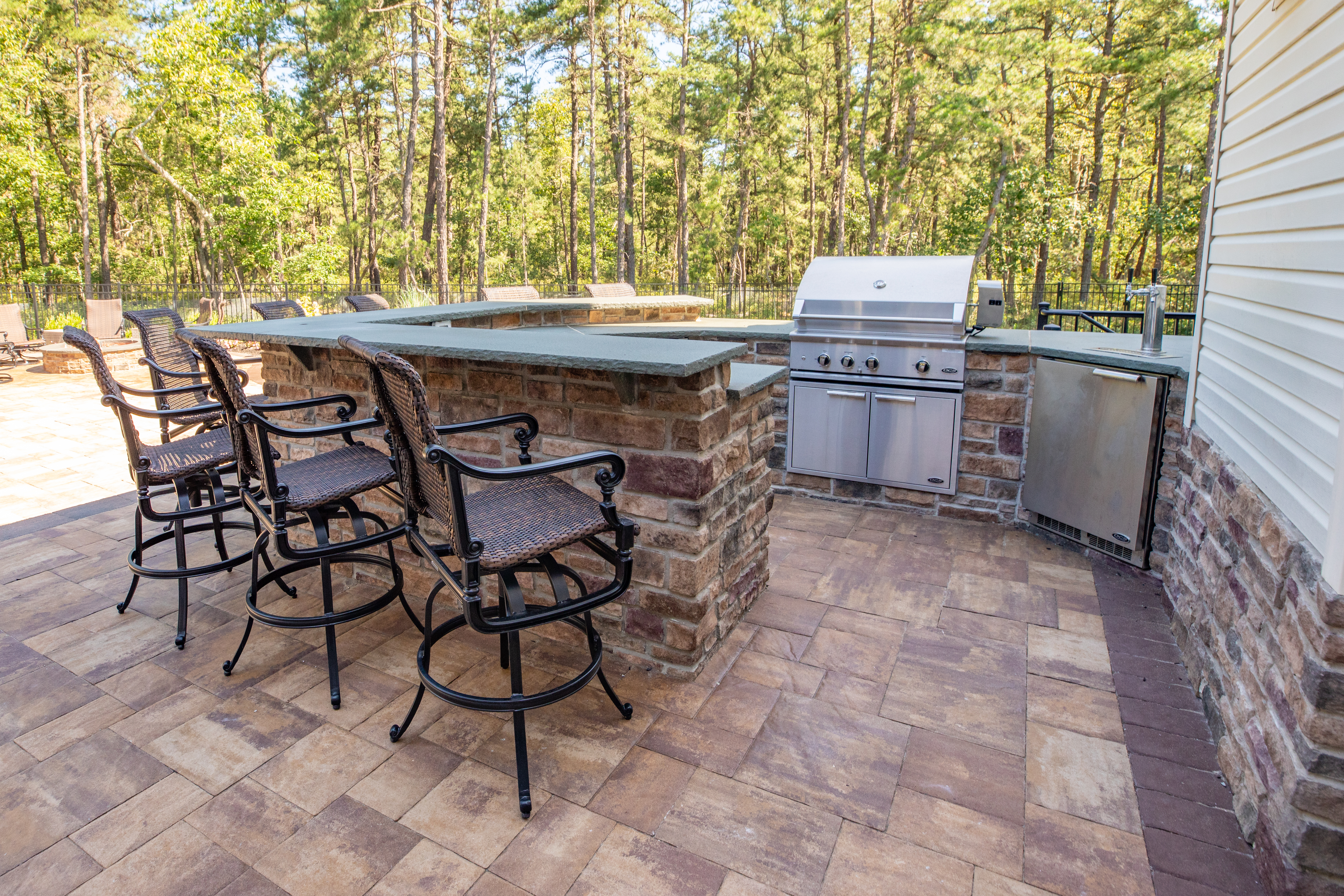 Outdoor kitchen featuring three bar stools, eating counter, stainless steel grill, and trees in the background. 