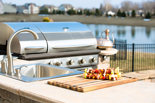 Closeup of stainless steel grill next to an outdoor sink and counter with a cutting board and kabobs, featuring a waterway.