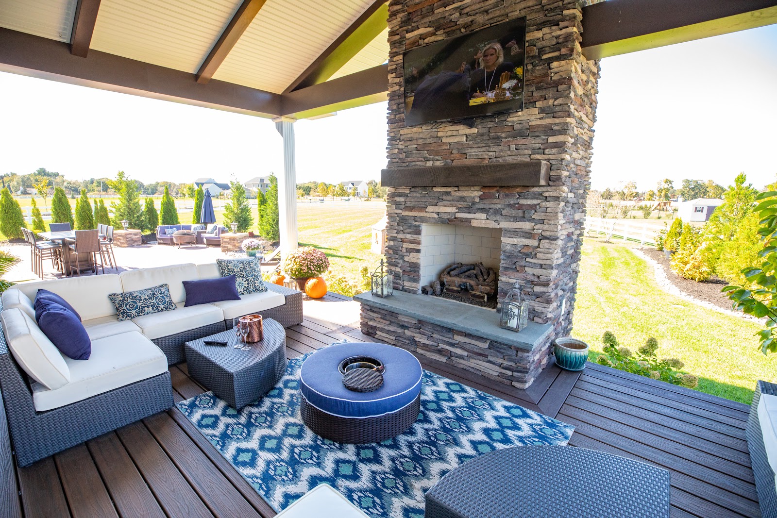 A backyard, wide-planked patio with stone-faced fireplace, blue-patterned rug, couch, and firepit surrounded with green landscape.
